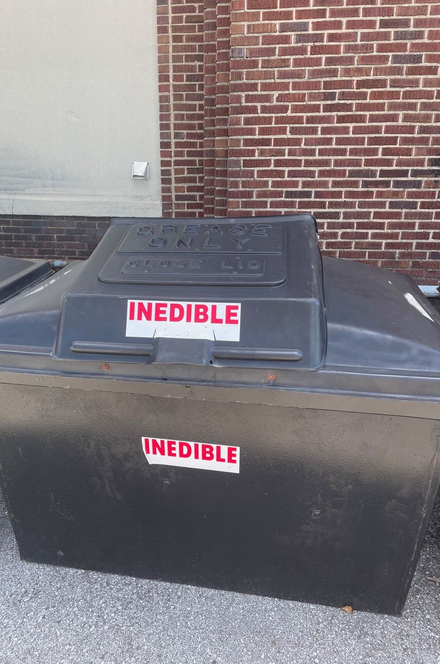 Black outdoor collection bin for used cooking oil against a brick wall